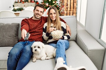 Young hispanic couple smiling happy watching movie sitting on the sofa with dog at home.