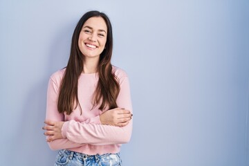 Young brunette woman standing over blue background happy face smiling with crossed arms looking at the camera. positive person.