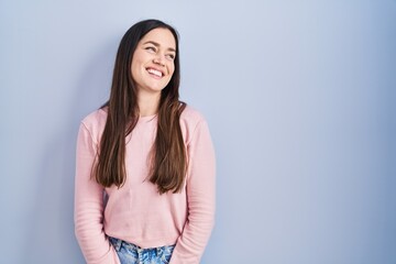 Obraz premium Young brunette woman standing over blue background looking away to side with smile on face, natural expression. laughing confident.