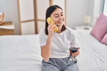 Fototapeta premium Young hispanic woman talking on the smartphone drinking wine at bedroom