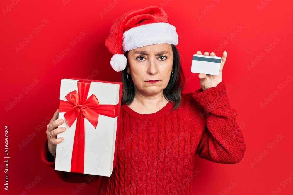 Wall mural middle age hispanic woman wearing christmas hat holding gift and credit card skeptic and nervous, fr