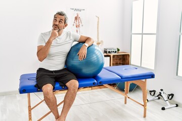 Middle age hispanic man at pain recovery clinic holding pilates ball with hand on chin thinking about question, pensive expression. smiling with thoughtful face. doubt concept.