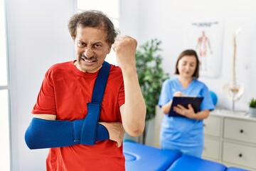 Southeast asian man wearing wearing arm on sling at rehabilitation clinic annoyed and frustrated shouting with anger, yelling crazy with anger and hand raised