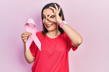 Obraz na płótnie Canvas Middle age hispanic woman holding pink cancer ribbon smiling happy doing ok sign with hand on eye looking through fingers