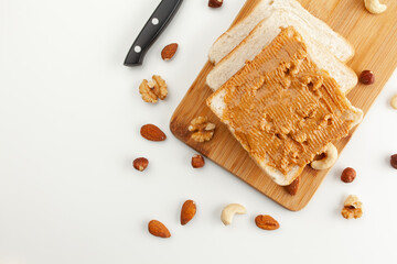 Square bread for toast with peanut butter on a wooden board. Nuts, a knife and a wooden cutting board with a sandwich and slices of bread on a white table.