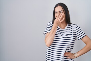 Young brunette woman wearing striped t shirt smelling something stinky and disgusting, intolerable smell, holding breath with fingers on nose. bad smell