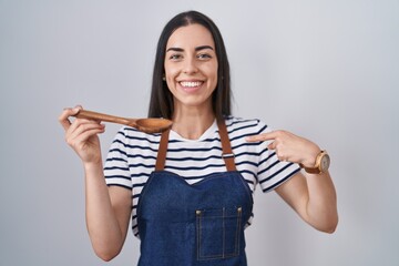 Young brunette woman wearing apron tasting food holding wooden spoon pointing finger to one self...