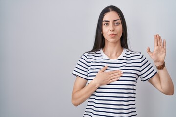 Young brunette woman wearing striped t shirt swearing with hand on chest and open palm, making a...