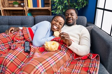 Young african american couple sitting on the sofa watching tv sticking tongue out happy with funny expression. - obrazy, fototapety, plakaty