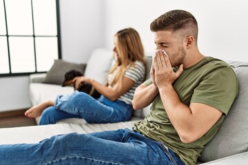 Young hispanic couple sitting on the sofa with dog at home. Man using handkerchief.