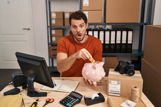 Young Hispanic Man Working At Small Business Ecommerce Holding Piggy Bank In Shock Face, Looking Skeptical And Sarcastic, Surprised With Open Mouth