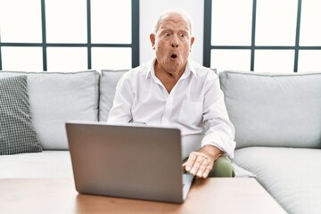 Senior man using laptop at home sitting on the sofa afraid and shocked with surprise expression, fear and excited face.