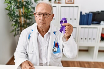Senior doctor man holding purple ribbon awareness thinking attitude and sober expression looking...