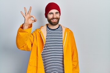 Fototapeta premium Caucasian man with beard wearing yellow raincoat smiling positive doing ok sign with hand and fingers. successful expression.