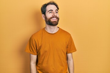 Caucasian man with beard wearing casual yellow t shirt looking away to side with smile on face,...