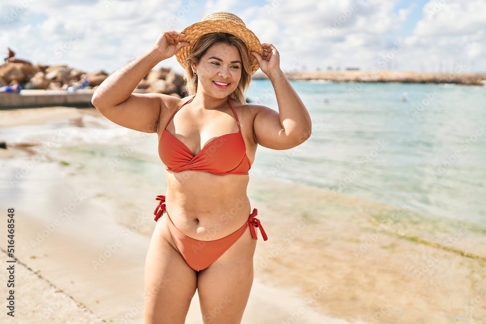 Wall mural young hispanic woman wearing bikini and summer hat standing at seaside