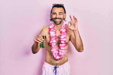 Young hispanic man wearing swimsuit and hawaiian lei drinking tropical cocktail smiling positive...