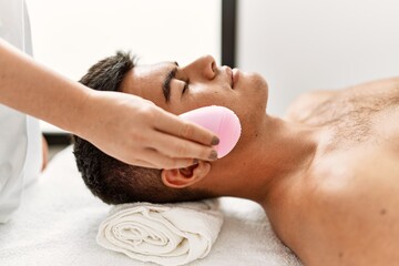 Young hispanic man relaxed having facial treatment cleaning face electric cleaner at beauty center