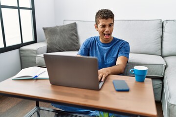 Young handsome hispanic man using laptop sitting on the floor winking looking at the camera with sexy expression, cheerful and happy face.