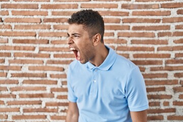 Brazilian young man standing over brick wall angry and mad screaming frustrated and furious, shouting with anger. rage and aggressive concept.