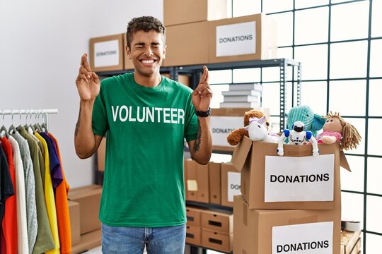 Young handsome hispanic man wearing volunteer t shirt at donations stand gesturing finger crossed smiling with hope and eyes closed. luck and superstitious concept.