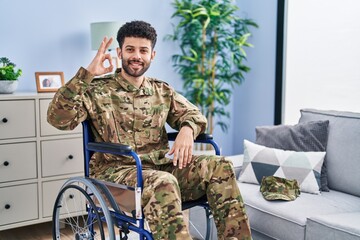 Fototapeta premium Arab man wearing camouflage army uniform sitting on wheelchair smiling positive doing ok sign with hand and fingers. successful expression.