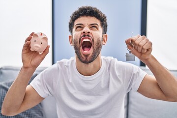 Arab man holding piggy bank and house keys angry and mad screaming frustrated and furious, shouting with anger looking up.
