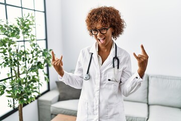 Young african american woman wearing doctor uniform and stethoscope shouting with crazy expression doing rock symbol with hands up. music star. heavy concept.