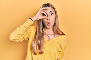 Beautiful hispanic woman wearing casual yellow sweater doing ok gesture shocked with surprised face, eye looking through fingers. unbelieving expression.