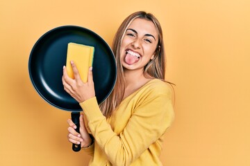 Beautiful hispanic woman cleaning cooking pan with scourer sticking tongue out happy with funny expression.