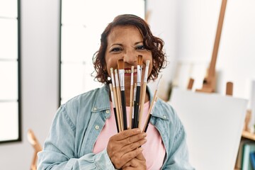 Middle age hispanic artist woman covering mouth with paintbrushes at art studio.