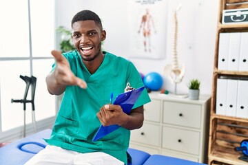 Young african american man working at pain recovery clinic smiling friendly offering handshake as...