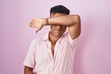 Young hispanic man standing over pink background covering eyes with arm, looking serious and sad....