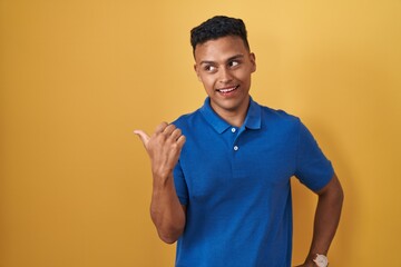 Young hispanic man standing over yellow background smiling with happy face looking and pointing to the side with thumb up.