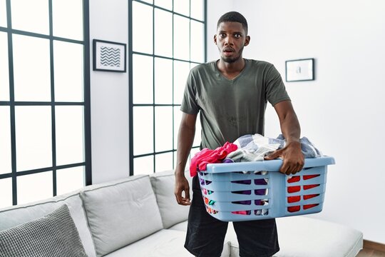 Young african american man doing laundry holding basket scared and amazed with open mouth for surprise, disbelief face