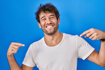 Hispanic young man standing over blue background looking confident with smile on face, pointing oneself with fingers proud and happy.