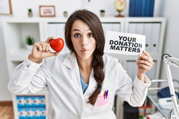 Young doctor woman holding your donation matters banner at the clinic making fish face with mouth...