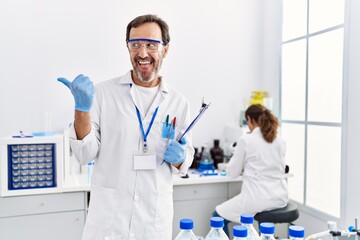 Middle age man working at scientist laboratory pointing thumb up to the side smiling happy with open mouth