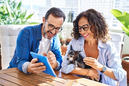 Middle age hispanic couple using touchpad sitting on table with dog at terrace