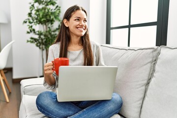 Young latin woman using laptop and drinking coffee sitting on sofa at home