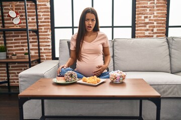 Young pregnant woman eating sweets at home scared and amazed with open mouth for surprise, disbelief face