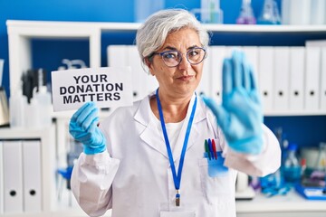 Middle age woman with grey hair working at scientist laboratory holding your donation matters banner with open hand doing stop sign with serious and confident expression, defense gesture
