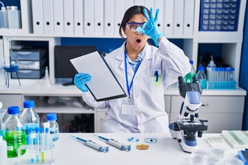 Fototapeta premium Hispanic young woman working at scientist laboratory doing ok gesture shocked with surprised face, eye looking through fingers. unbelieving expression.
