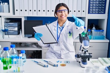 Hispanic young woman working at scientist laboratory approving doing positive gesture with hand, thumbs up smiling and happy for success. winner gesture.