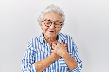 Senior woman with grey hair standing over white background smiling with hands on chest with closed...