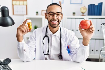 African american doctor man holding prescription pills and fresh apple winking looking at the camera with sexy expression, cheerful and happy face.