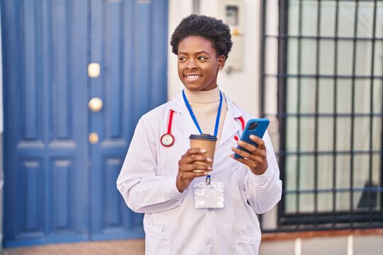 African american woman wearing doctor uniform using smartphone drinking coffee at street