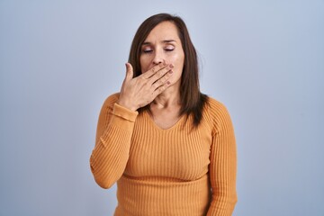 Middle age brunette woman standing wearing orange sweater bored yawning tired covering mouth with hand. restless and sleepiness.