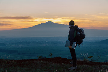 Unrecognizable young man with a backpack holding a laptop with a mountain in the background