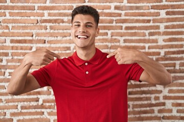 Young hispanic man standing over bricks wall looking confident with smile on face, pointing oneself...